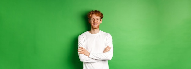 Бесплатное фото Молодой амбициозный мужчина с рыжими волосами стоит на зеленом фоне, держась за руки, скрещенные на груди и