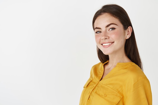 Young ambitious woman looking at front, smiling confident, standing over white wall in yellow blouse