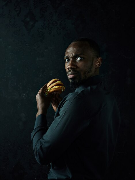 young american man eating hamburger and looking away on black studio background