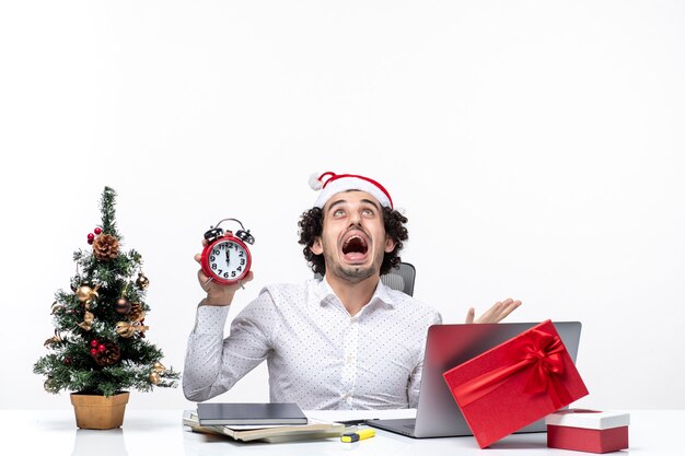 Young angry business person with santa claus hat and holding clock and sitting in the office on dark background