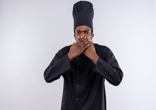 Young annoyed afro-american cook in chef uniform closes mouth with both hands isolated on white background with copy space