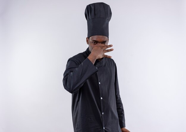Young annoyed afro-american cook in chef uniform closes nose with hand on white  with copy space