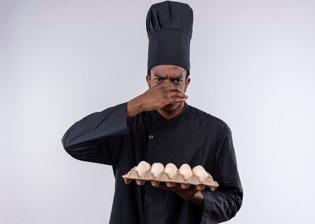 Young annoyed afro-american cook in chef uniform holds batch of eggs and closes nose with hand isolated on white background with copy space