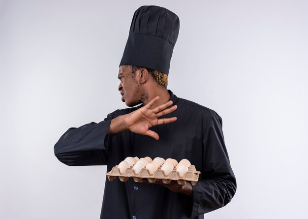 Young annoyed afro-american cook in chef uniform holds batch of eggs and gestures go away hand sign isolated on white background with copy space