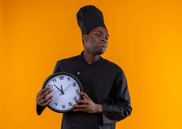 シェフの制服を着た若いイライラしたアフリカ系アメリカ人の料理人は時計を保持し、コピースペースでオレンジ色の側面を見ます