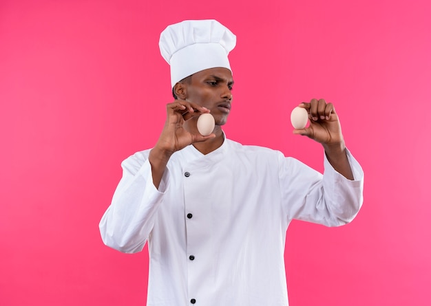 Free photo young annoyed afro-american cook in chef uniform holds eggs in both hands isolated on pink background with copy space