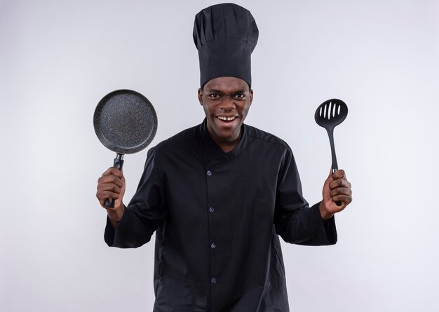 Young annoyed afro-american cook in chef uniform holds frying pan and spatula on white  with copy space