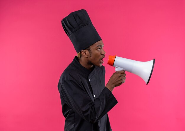 요리사 제복을 입은 젊은 화가 아프리카 계 미국인 요리사는 복사 공간이 분홍색 배경에 고립 된 시끄러운 스피커를 보유하고 있습니다.