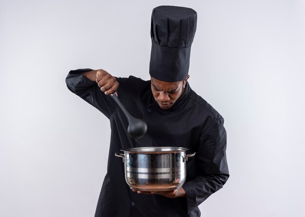 Young annoyed afro-american cook in chef uniform holds saucepan and spoon isolated on white background with copy space