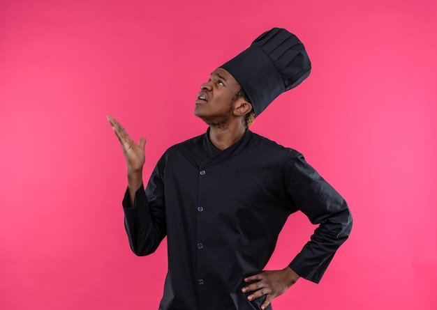 Молодой раздраженный афро-американский повар в униформе шеф-повара показывает рукой, изолированной на розовом фоне с копией пространства