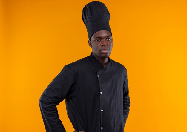 Young annoyed afro-american cook in chef uniform puts hands on waist and looks at camera isolated on orange background with copy space