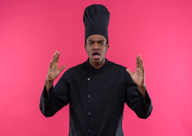 요리사 제복을 입은 젊은 화가 아프리카 계 미국인 요리사가 복사 공간이 분홍색 배경에 고립 된 손을 올립니다.