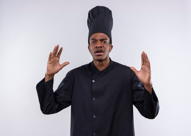 Young annoyed afro-american cook in chef uniform raises hands up isolated on white background with copy space