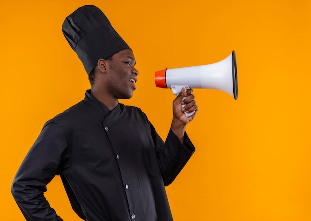 Young annoyed afro-american cook in chef uniform stands sideways and speaks through loud speaker on orange with copy space