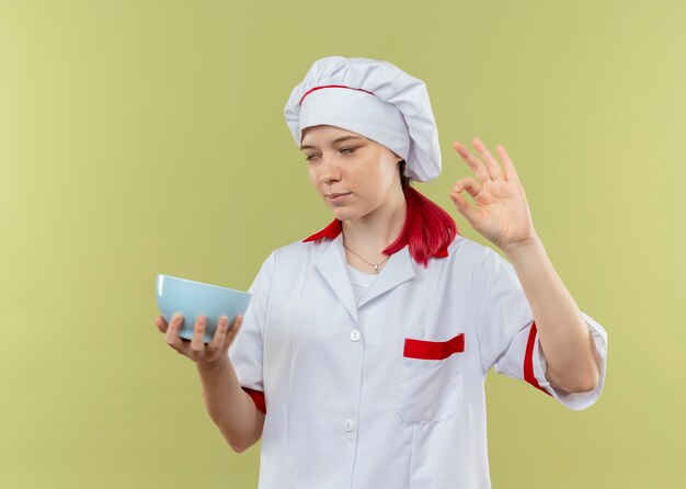 Young annoyed blonde female chef in chef uniform holds bowl and gestures ok hand sign rolling eyes isolated on green wall