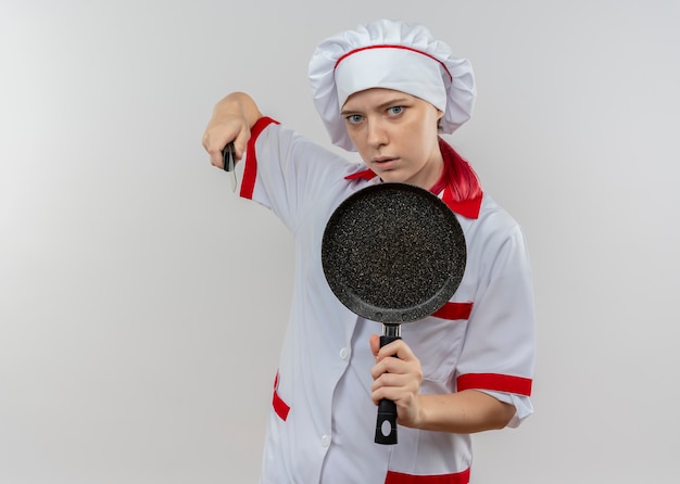 Il giovane chef femminile biondo infastidito in uniforme del cuoco unico tiene la padella e il coltello isolati sulla parete bianca