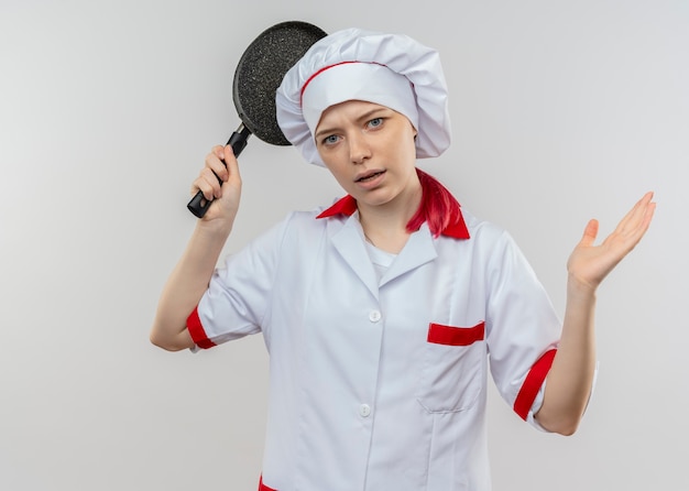 Il giovane chef femminile biondo infastidito in uniforme del cuoco unico tiene la padella e sembra isolato sulla parete bianca