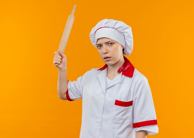 Young annoyed blonde female chef in chef uniform holds rolling pin and looks isolated on orange wall