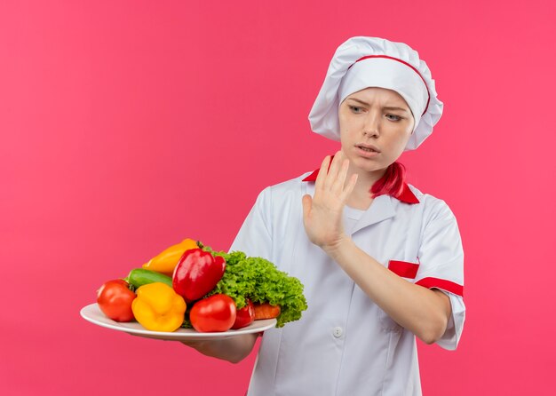 요리사 유니폼에 젊은 화가 금발 여성 요리사는 접시에 야채를 보유하고 분홍색 벽에 고립 손으로 밀어 척