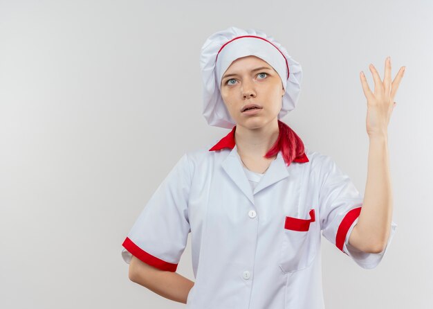 요리사 제복을 입은 젊은 화가 금발 여성 요리사가 외모와 흰 벽에 고립 된 손을 올립니다.