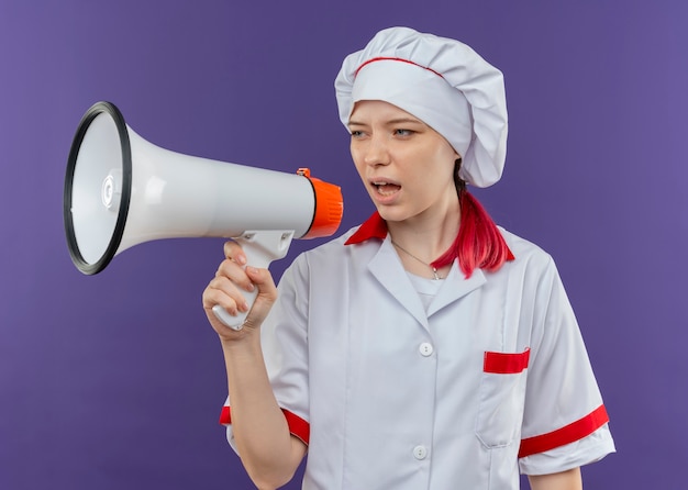 Young annoyed blonde female chef in chef uniform shouts through loud speaker isolated on violet wall