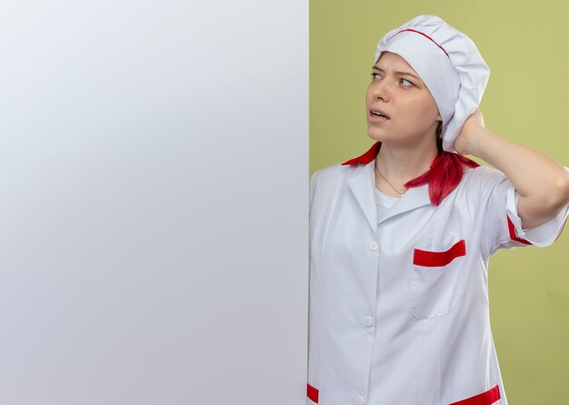 Young annoyed blonde female chef in chef uniform stands behind and looks at white wall isolated on green wall