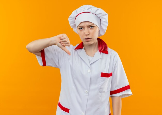 Giovane chef femmina bionda infastidito in uniforme da chef pollici giù isolato sulla parete arancione