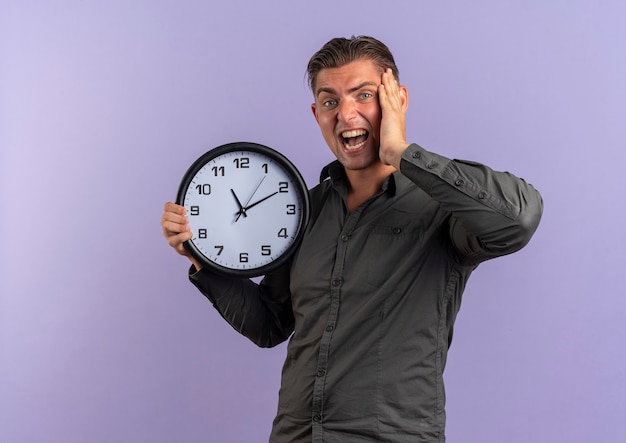 Young annoyed blonde handsome man holds clock and puts hand on face isolated on violet background with copy space