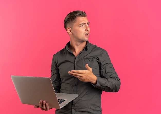 Young annoyed blonde handsome man holds and points at laptop looking at side isolated on pink background with copy space