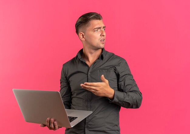 若いイライラする金髪のハンサムな男は、コピースペースでピンクの背景に分離された側を見てラップトップを保持し、指さします