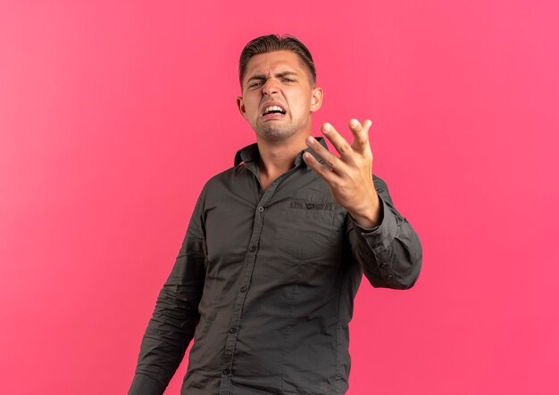 Foto gratuita il giovane uomo bello biondo infastidito esamina la macchina fotografica con la mano alzata isolata su fondo rosa con lo spazio della copia