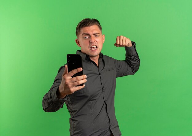 Young annoyed blonde handsome man looks at phone and keeps fist up ready to punch isolated on green background with copy space