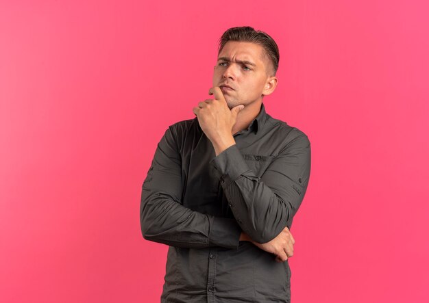 Young annoyed blonde handsome man puts hand on chin isolated on pink background with copy space