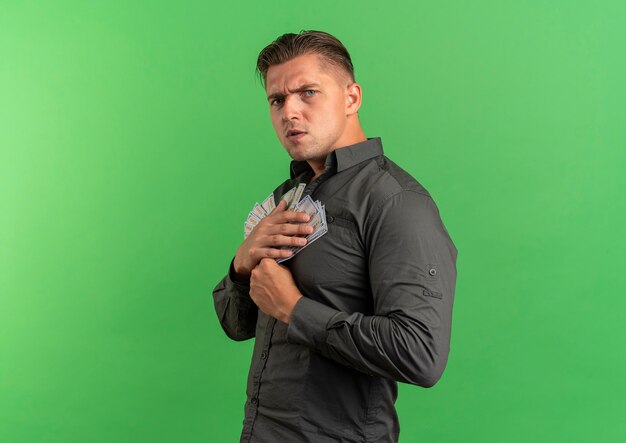 Free photo young annoyed blonde handsome man stands sideways holds money looking at camera isolated on green background with copy space