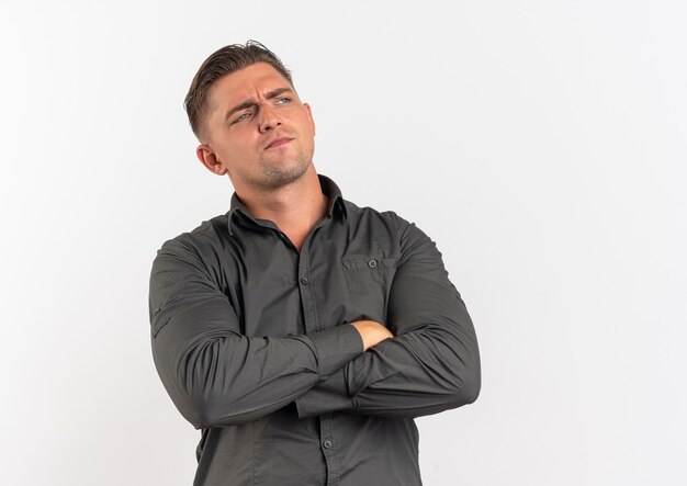 Young annoyed blonde handsome man stands with crossed arms looking at side isolated on white background with copy space