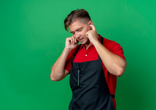 Young annoyed blonde male barber in uniform blockes ears with fingers isolated on green space with copy space