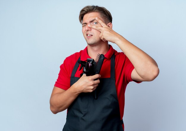 Young annoyed blonde male barber in uniform holds barber tools looking through fingers isolated on white space with copy space