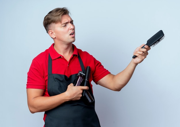 Young annoyed blonde male barber in uniform holds barber tools pointing at side with comb isolated on white space with copy space