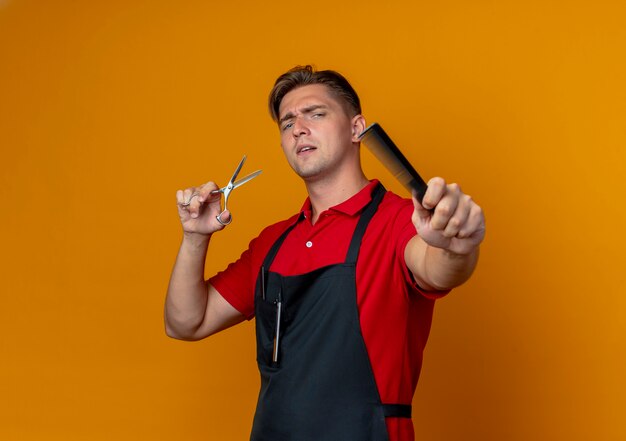 Young annoyed blonde male barber in uniform holds comb and scissors isolated on orange space with copy space