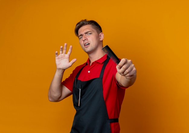 Young annoyed blonde male barber in uniform holds comb stretching out hands isolated on orange background with copy space