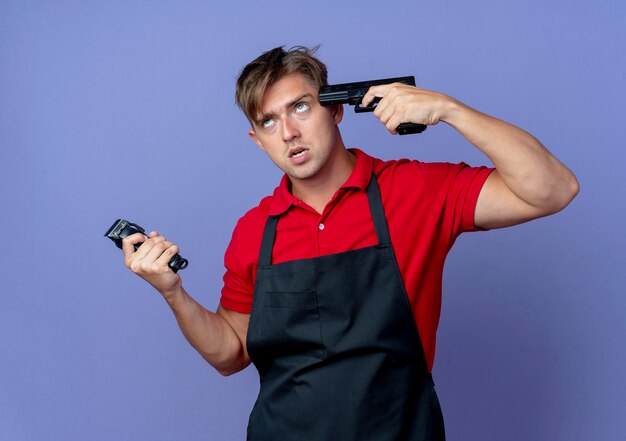 Young annoyed blonde male barber in uniform holds gun to temple looking up holding hair clipper isolated on violet space with copy space