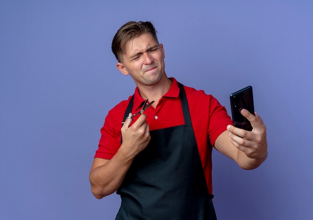 Молодой раздраженный светловолосый парикмахер в униформе держит ножницы, глядя на телефон, изолированный на фиолетовом пространстве с копией пространства
