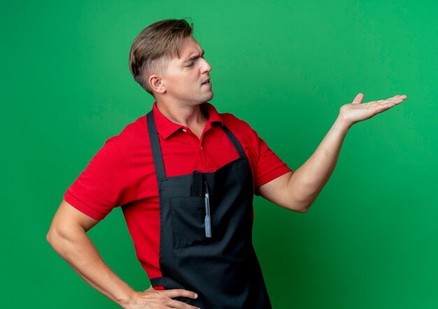 Young annoyed blonde male barber in uniform looks and points at side with hand isolated on green space with copy space