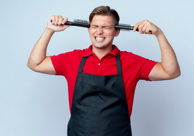 Young annoyed blonde male barber in uniform puts combs on temples isolated on white space with copy space