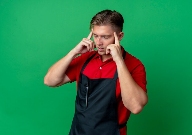 Young annoyed blonde male barber in uniform puts fingers on temple isolated on green space with copy space