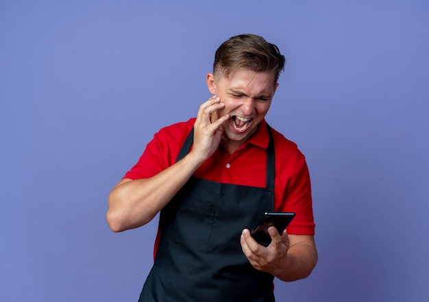 Young annoyed blonde male barber in uniform puts hand on face looking at phone isolated on violet space with copy space