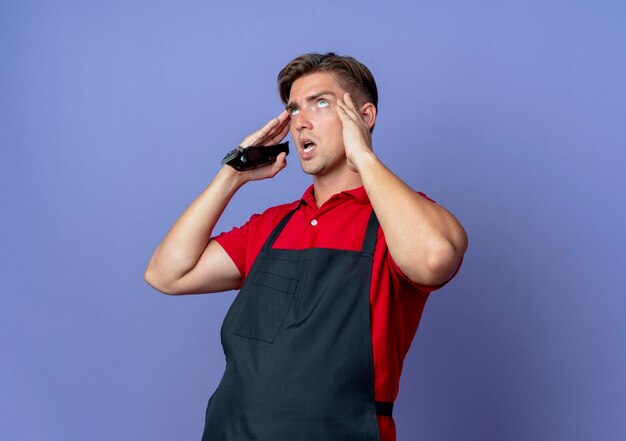 Young annoyed blonde male barber in uniform puts hands on temples holding hair clipper isolated on violet space with copy space