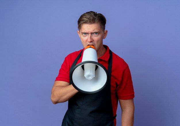 Young annoyed blonde male barber in uniform shouts into loud speaker isolated on violet space with copy space