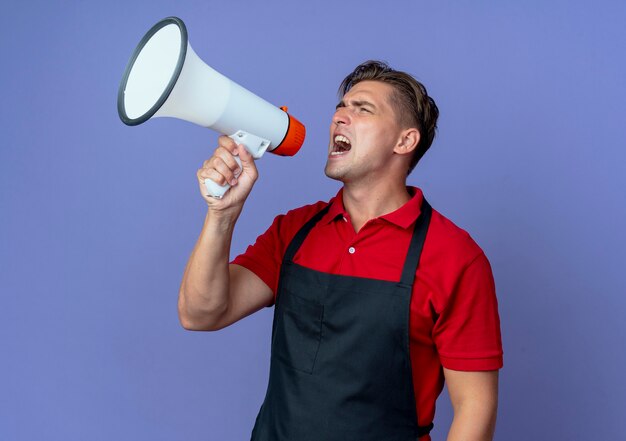 Young annoyed blonde male barber in uniform shouts into loud speaker looking at side isolated on violet space with copy space