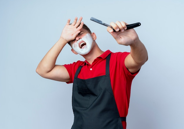 Young annoyed blonde male barber in uniform smeared face with shaving foam holding straight razor putting hand on forehead isolated on white space with copy space